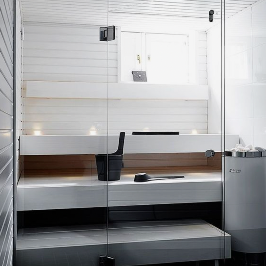 Sleek and contemporary, the sauna features white wooden walls and dark benches, with a white sauna heater to the side, complemented by a black bucket and ladle.